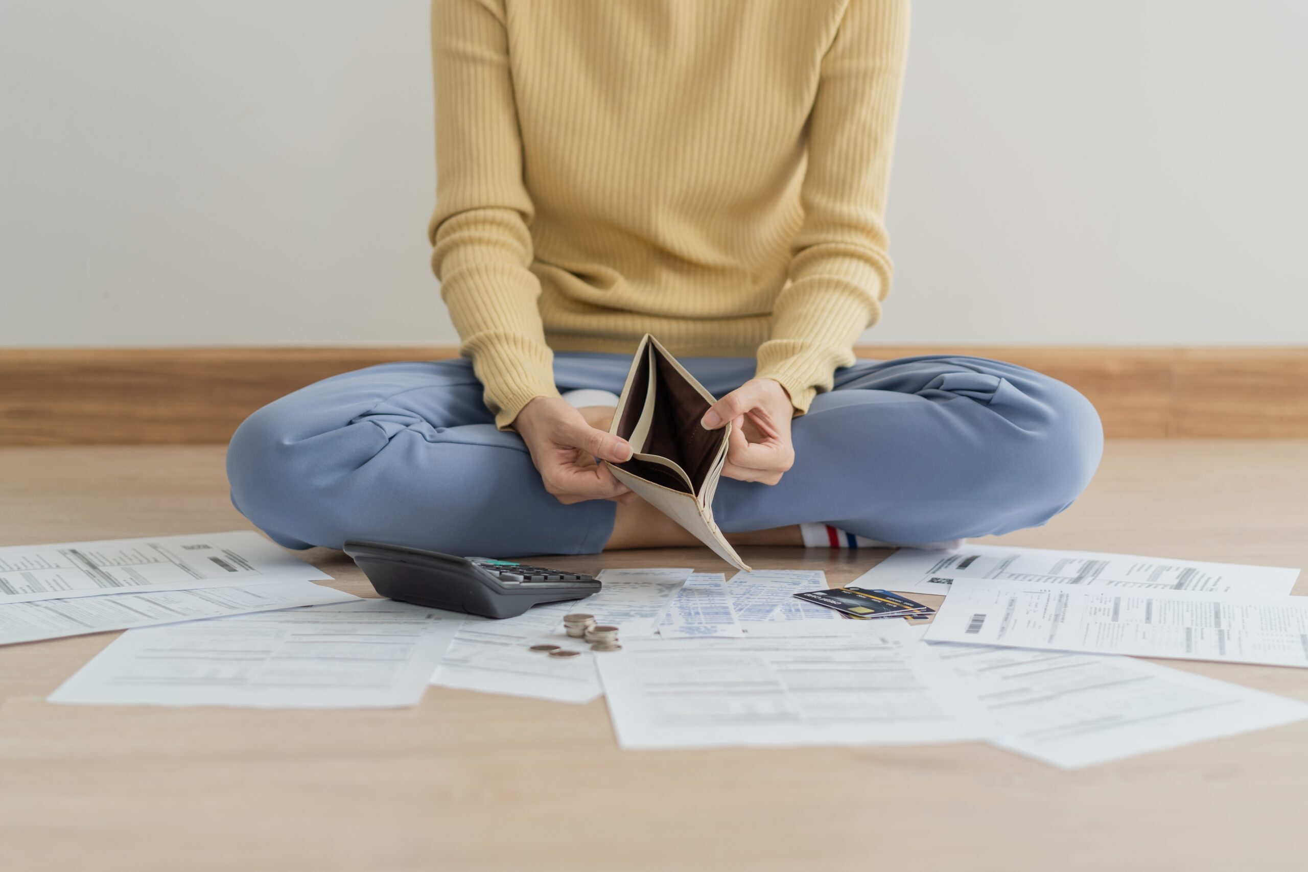 Woman Holding An Empty Wallet While Looking At A Pile Of Paperwork And A Calculator.